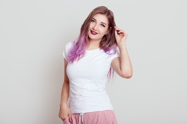 Mulher jovem e atraente com lábios brilhantes e cabelo lilás, em pé e tocando seu cabelo, expressando emoções positivas, posando isolado sobre uma parede cinza.