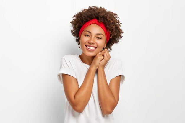 Mulher jovem e atraente com penteado afro, mantém as mãos juntas, usa bandana vermelha e roupas casuais