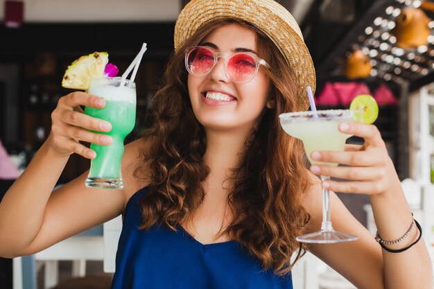 Mulher jovem e atraente com vestido azul e chapéu de palha usando óculos de sol rosa, bebendo coquetéis de álcool nas férias tropicais e sentada à mesa no bar