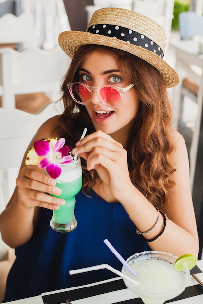 Mulher jovem e atraente com vestido azul e chapéu de palha usando óculos de sol rosa, bebendo coquetéis de álcool nas férias tropicais e sentada à mesa no bar
