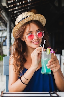 Mulher jovem e atraente com vestido azul e chapéu de palha usando óculos de sol rosa, bebendo coquetéis de álcool nas férias tropicais, sentada à mesa em um bar com roupa estilo verão, sorrindo feliz em clima de festa