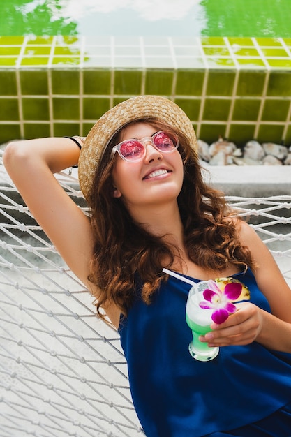 Mulher jovem e atraente com vestido azul e chapéu de palha usando óculos de sol rosa, bebendo um coquetel de álcool nas férias, sentada na rede com roupa estilo verão, sorrindo feliz em clima de festa