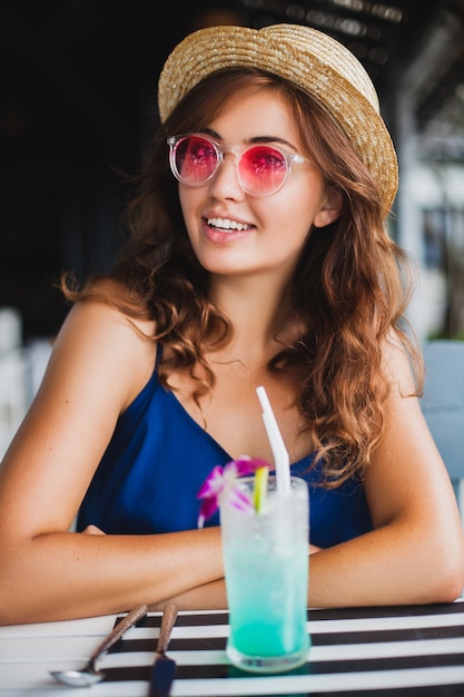 Mulher jovem e atraente com vestido azul e chapéu de palha usando óculos de sol rosa, bebendo um coquetel de álcool nas férias tropicais e sentada à mesa no bar