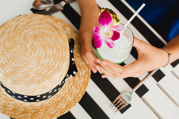 Mulher jovem e atraente com vestido azul e chapéu de palha usando óculos de sol rosa, bebendo um coquetel de álcool nas férias tropicais e sentada à mesa no bar