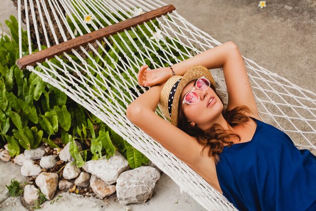 Mulher jovem e atraente com vestido azul e chapéu de palha usando óculos de sol rosa relaxando nas férias deitada na rede com roupa estilo verão, sorrindo feliz