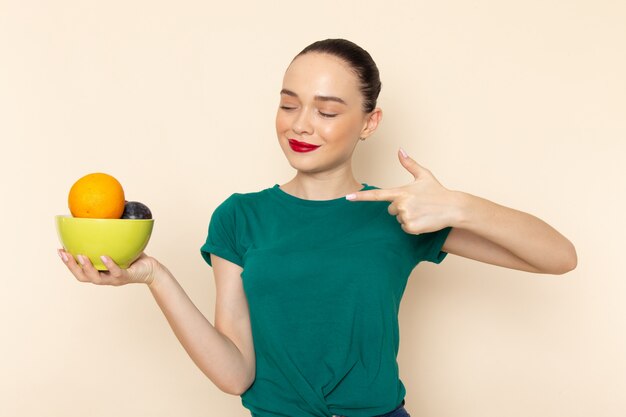 Mulher jovem e atraente de frente com camisa verde escura segurando um prato com frutas apontando