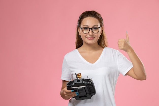 Mulher jovem e atraente de frente, em smi de camiseta branca e segurando o controle remoto no fundo rosa