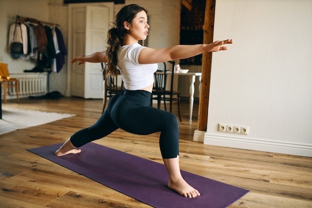 Mulher jovem e atraente descalça praticando ioga em casa, em pé no tapete, fazendo pose de guerreiro II ou Virabhadrasana, fortalecendo pernas, abrindo quadris e desenvolvendo concentração e equilíbrio
