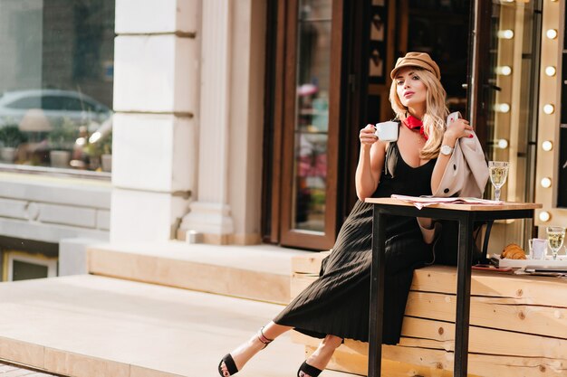 Mulher jovem e atraente descansando depois do trabalho no café favorito e apreciando o sabor do café. Retrato ao ar livre de uma menina loira com roupa elegante e relaxante no fim de semana.