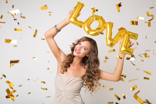 Mulher jovem e atraente e elegante comemorando o ano novo, segurando balões de ar, cartas de amor, confetes dourados voando, sorrindo, feliz, isolada, usando vestido de festa