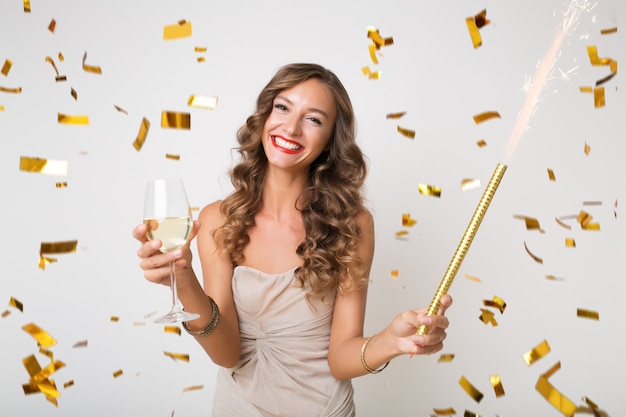 Mulher jovem e atraente elegante comemorando ano novo, bebendo champanhe, confete dourado voando, sorrindo feliz, isolada, usando vestido de festa