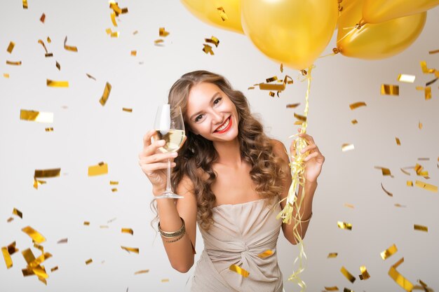 Mulher jovem e atraente elegante comemorando ano novo, bebendo champanhe segurando balões de ar, confete dourado voando, sorrindo feliz, branco, isolado, usando vestido de festa