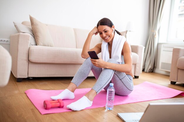 Mulher jovem e atraente fazendo uma pausa enquanto se exercita em casa Mulher usando telefone inteligente no colchonete na frente de seu laptop