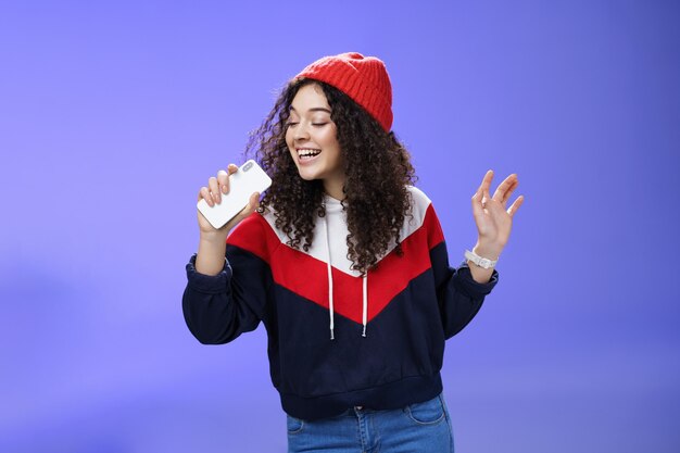 Mulher jovem e atraente feliz com cabelo encaracolado no chapéu cantando, aproveitando o dia de inverno perfeito cantando no smartphone, segurando o celular como um microfone, adora karaokê sobre fundo azul.