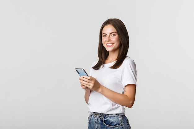 Mulher jovem e atraente morena usando telefone celular e sorrindo para a câmera.