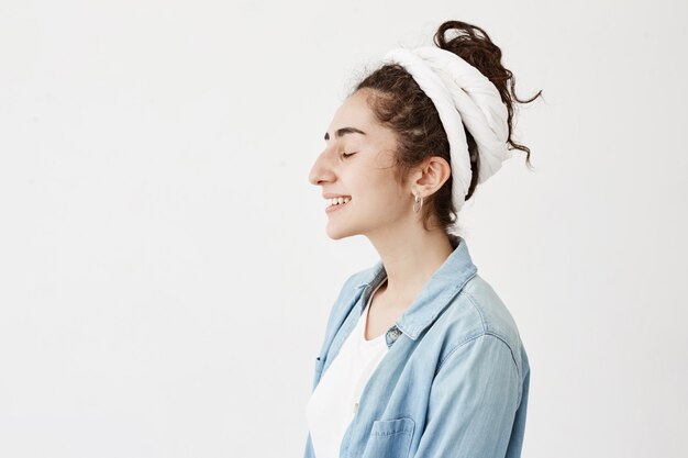 Mulher jovem e atraente na camisa de pano e jeans, relaxante dentro de casa, com sorriso bonito e olhos fechados, posando contra parede branca com espaço de cópia para o seu texto ou conteúdo publicitário