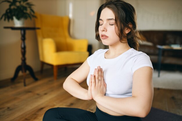 Mulher jovem e atraente praticando meditação na sala de estar para atingir um estado mental calmo e estável, mantendo os olhos fechados.