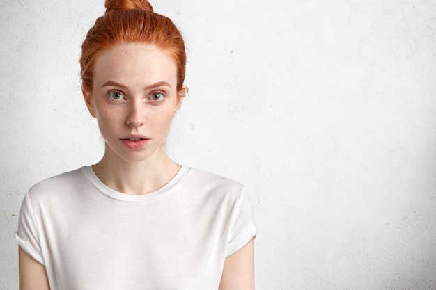 Mulher jovem e atraente ruiva com expressão séria, pele sardenta, vestida casualmente, posa contra o espaço em branco