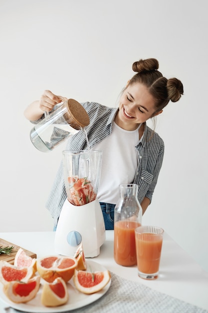 Foto grátis mulher jovem e atraente sorrindo adicionando água no liquidificador com pedaços de toranja e alecrim. nutrição alimentar de dieta saudável.