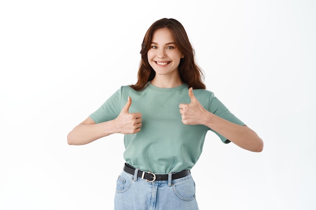 Foto grátis mulher jovem e atraente sorrindo e mostrando os polegares em aprovação, gostar e recomendar coisas boas, elogiar o bom trabalho, gesto bem executado, dizer sim, em pé contra um fundo branco