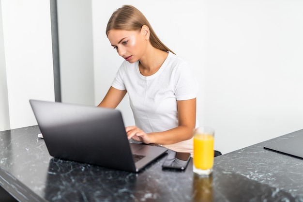 Mulher jovem e atraente trabalhando em um laptop e tomando suco enquanto está sentado na cozinha