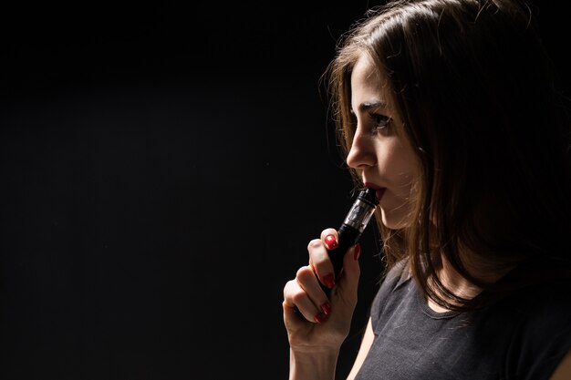 Mulher jovem e atraente vaping e soprando fumaça isolada na parede preta