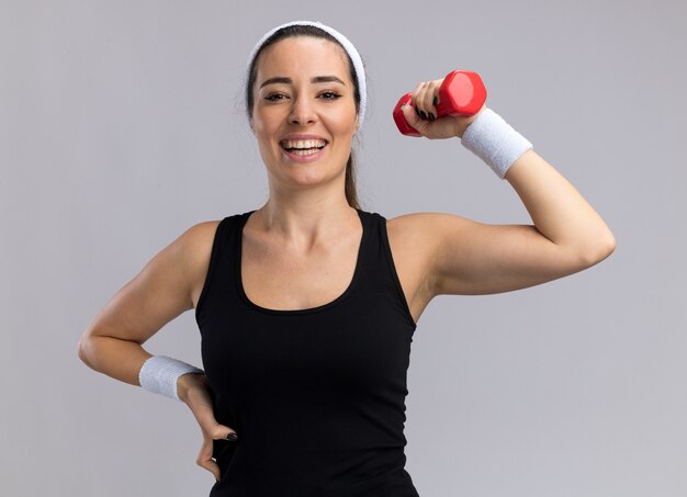 Mulher jovem e bonita, alegre e esportiva, usando fita para a cabeça e pulseiras, olhando para a frente, mantendo a mão na cintura, levantando halteres isolado na parede branca