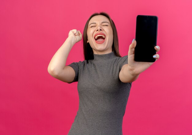 Mulher jovem e bonita alegre esticando o celular para a câmera levantando o punho com os olhos fechados, isolado no fundo rosa com espaço de cópia