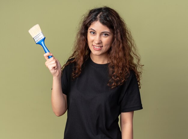 Mulher jovem e bonita alegre segurando um pincel, olhando para frente, isolada na parede verde oliva com espaço de cópia