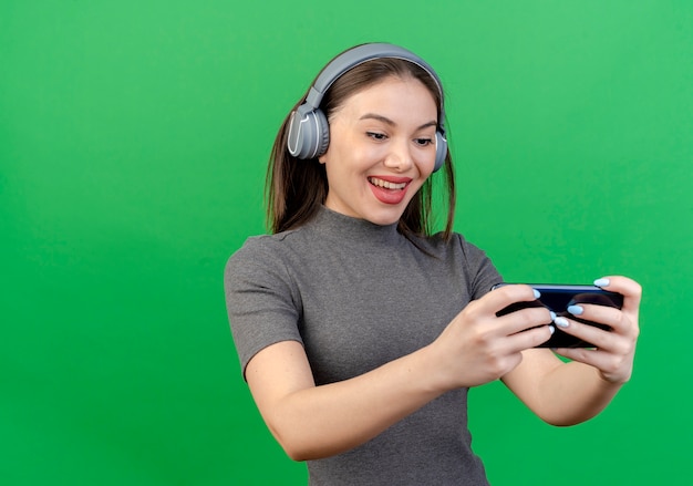 Mulher jovem e bonita alegre usando fones de ouvido e telefone celular isolado em um fundo verde com espaço de cópia