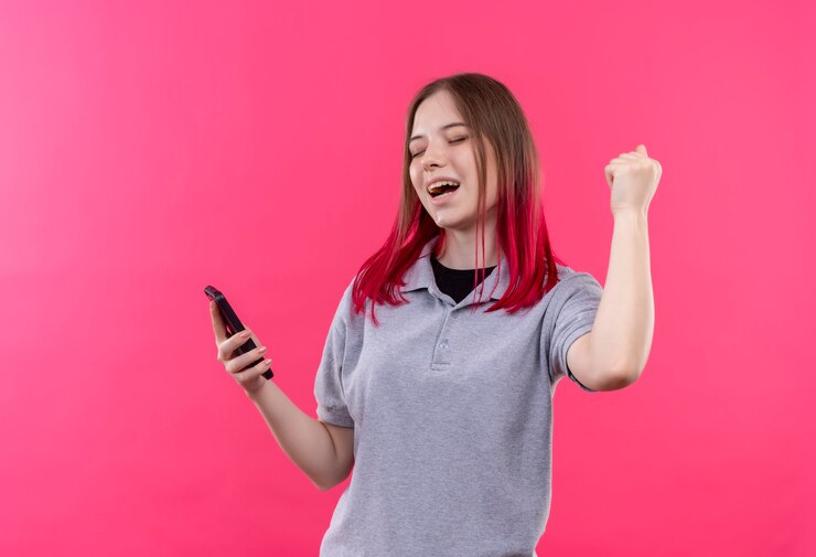 Foto grátis mulher jovem e bonita alegre vestindo uma camiseta cinza segurando um telefone e mostrando um gesto de sim na parede rosa isolada