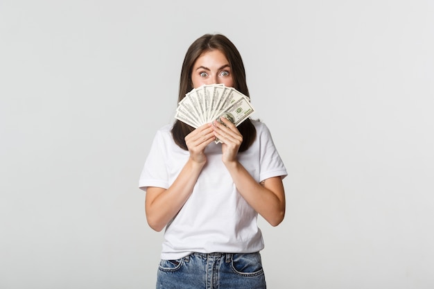 Mulher jovem e bonita animada segurando dinheiro sobre o rosto, branco de pé.