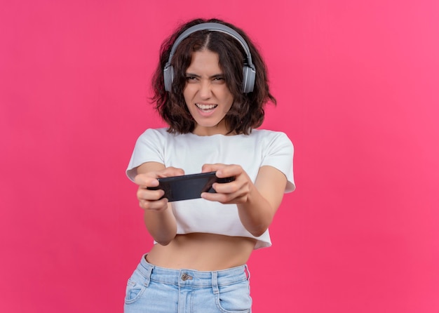 Foto grátis mulher jovem e bonita animada usando fones de ouvido e segurando o celular na parede rosa com espaço de cópia