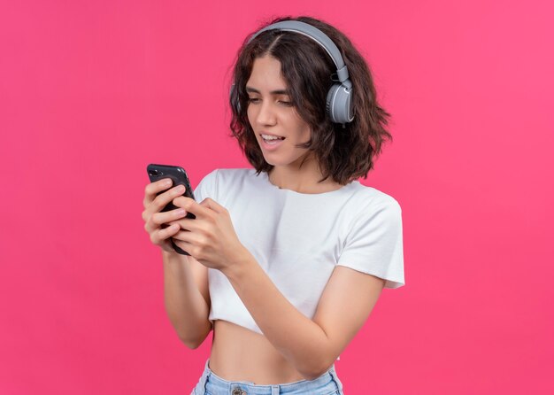 Mulher jovem e bonita animada usando fones de ouvido e segurando o celular na parede rosa com espaço de cópia