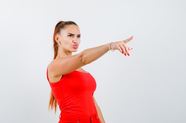 Foto grátis mulher jovem e bonita apontando para longe, fazendo beicinho com os lábios em um top vermelho e olhando a vista frontal, focada.