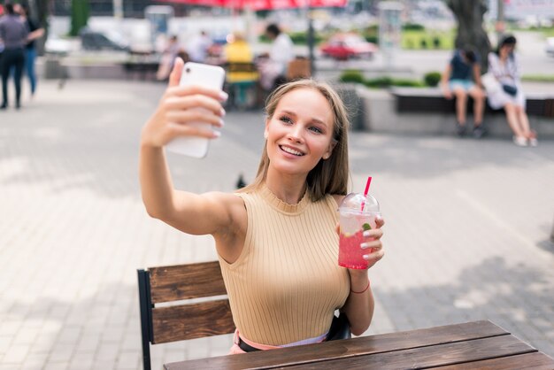 Mulher jovem e bonita bebendo mojito fazendo selfie no café da rua de verão