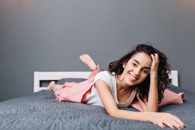 Mulher jovem e bonita bonita de pijama, deitada na cama em um apartamento moderno. Sorrindo, relaxando em casa pela manhã, expressando positividade