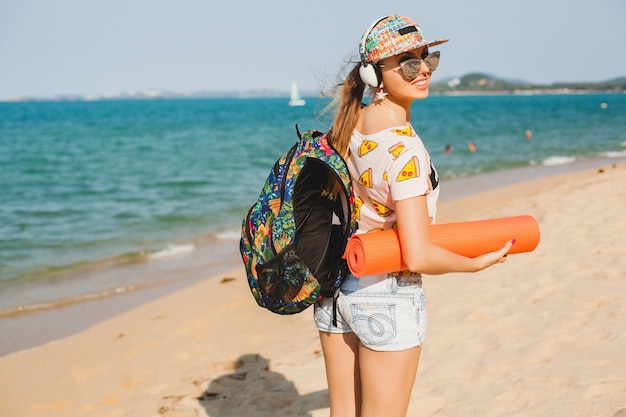 Mulher jovem e bonita caminhando na praia com tapete de ioga, ouvindo música em fones de ouvido, estilo moderno estilo swag, shorts jeans, camiseta, mochila, boné, óculos de sol, ensolarado, fim de semana de verão, alegre