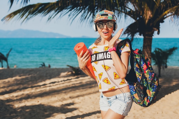 Foto grátis mulher jovem e bonita caminhando na praia com tapete de ioga, ouvindo música em fones de ouvido, estilo moderno estilo swag, shorts jeans, camiseta, mochila, boné, óculos de sol, ensolarado, fim de semana de verão, alegre