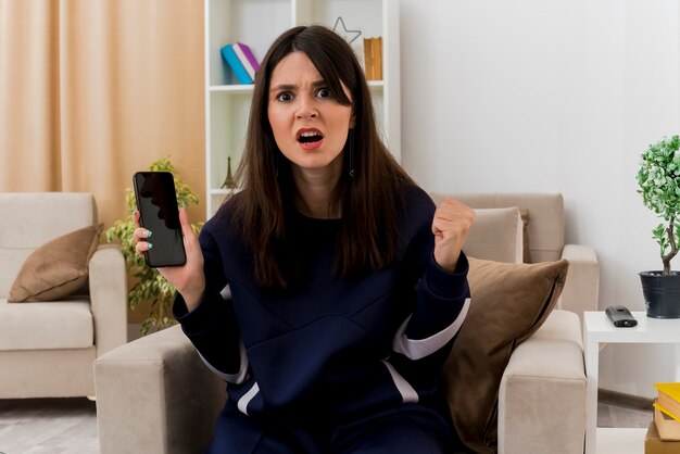 Mulher jovem e bonita caucasiana com raiva sentada em uma poltrona em uma sala projetada segurando um telefone celular, olhando e cerrando o punho