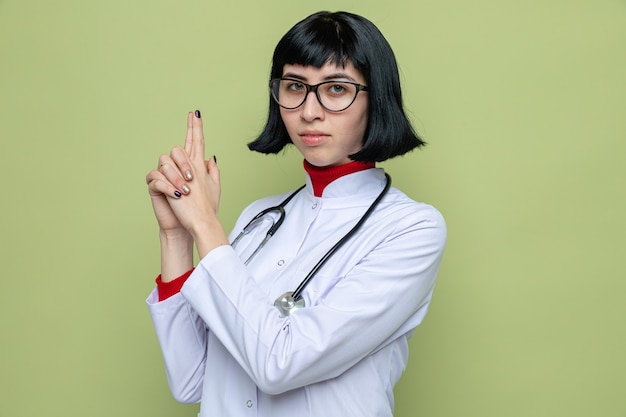 Mulher jovem e bonita caucasiana confiante com óculos em uniforme de médico com estetoscópio apontando uma arma com as duas mãos