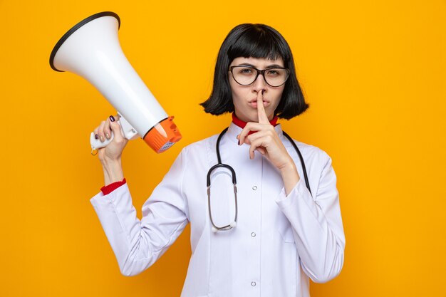 Mulher jovem e bonita caucasiana confiante com óculos em uniforme de médico com estetoscópio segurando um alto-falante e fazendo gesto de silêncio