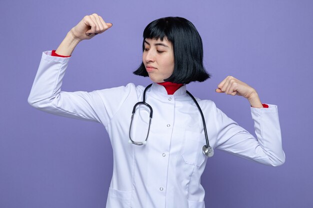 Mulher jovem e bonita caucasiana confiante em uniforme de médico com estetoscópio tensiona bíceps olhando para o lado