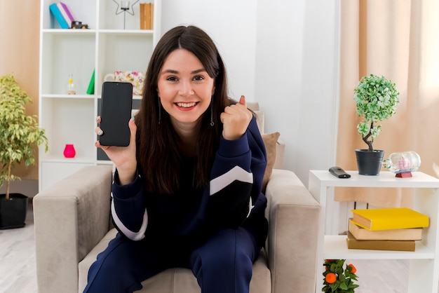 Mulher jovem e bonita caucasiana feliz sentada na poltrona na sala projetada, mostrando o telefone celular e fazendo o gesto de sim