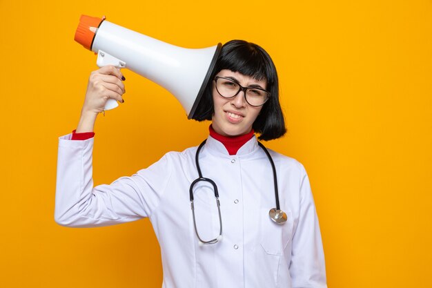 Mulher jovem e bonita caucasiana insatisfeita com óculos em uniforme de médico com estetoscópio segurando um alto-falante