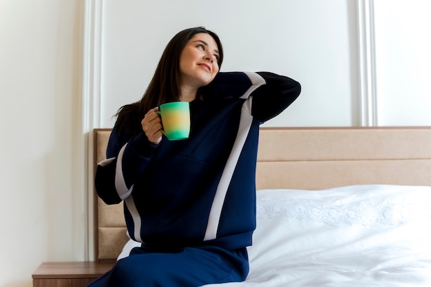 Mulher jovem e bonita caucasiana satisfeita sentada na cama no quarto segurando um copo, olhando para o lado, colocando a mão atrás do pescoço