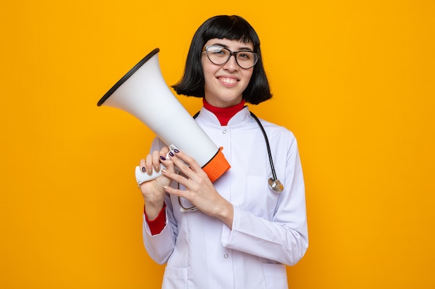 Mulher jovem e bonita caucasiana sorridente com óculos ópticos em uniforme de médico com estetoscópio segurando um alto-falante