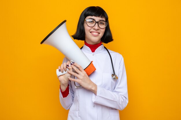 Mulher jovem e bonita caucasiana sorridente com óculos ópticos em uniforme de médico com estetoscópio segurando um alto-falante