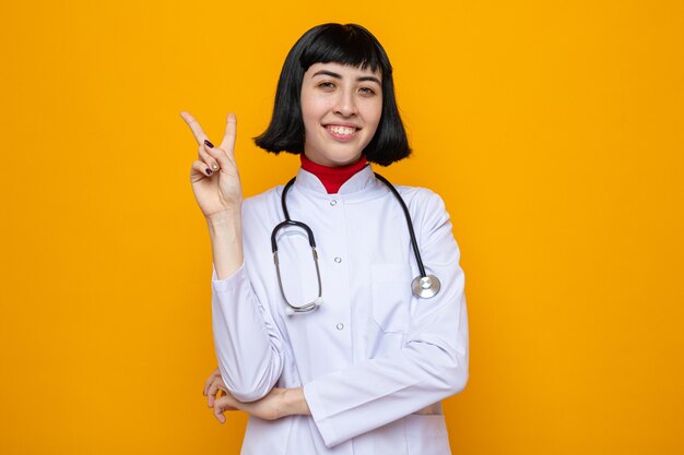 Mulher jovem e bonita caucasiana sorridente com uniforme de médico e estetoscópio gesticulando sinal de vitória