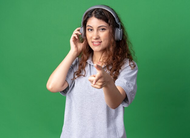 Foto grátis mulher jovem e bonita caucasiana sorridente usando fones de ouvido, olhando e apontando para a frente tocando fones de ouvido